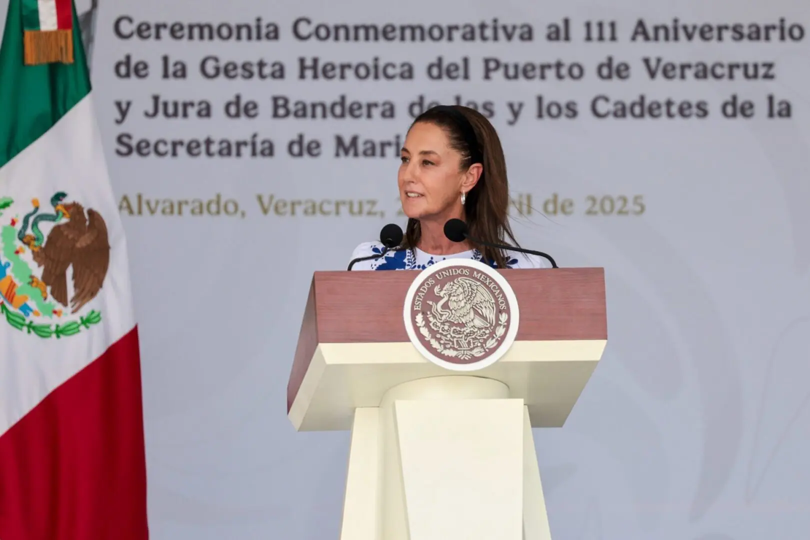 Woman speaking at podium with Mexican flag.