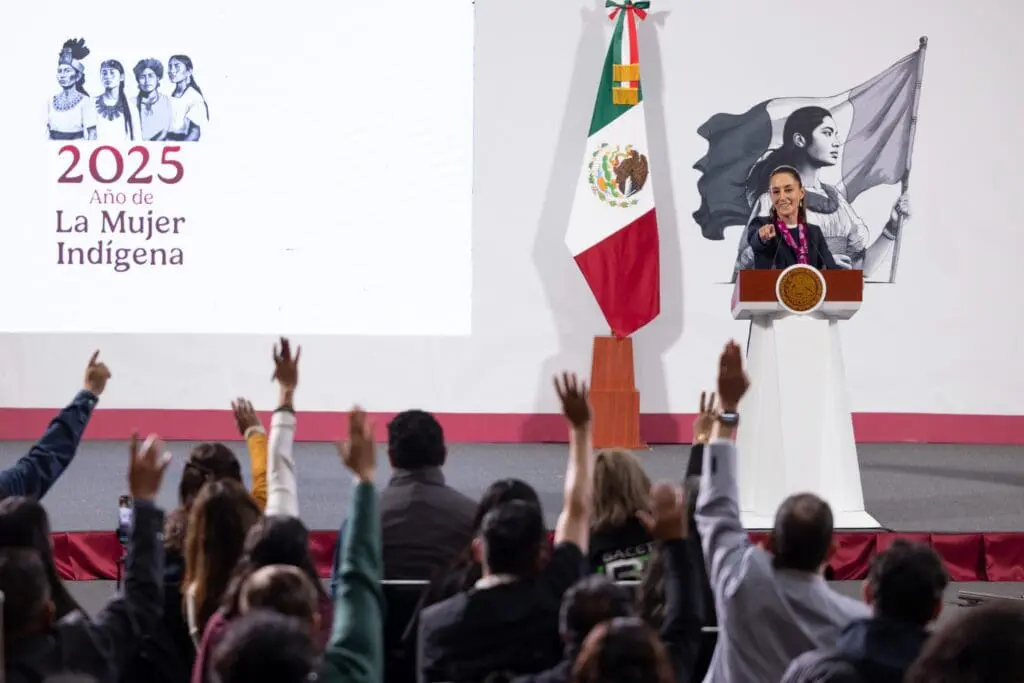 A speaker at a podium with the Mexican flag and an audience raising hands.