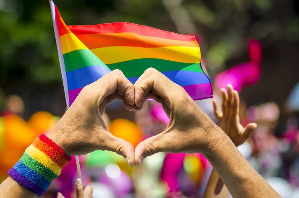 Hands forming heart, rainbow flag background.