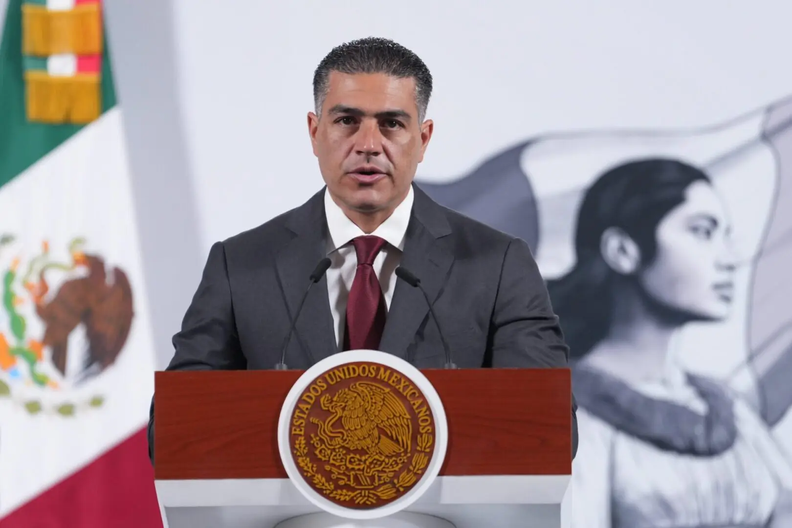 A man in a suit speaking at a podium with an emblem.
