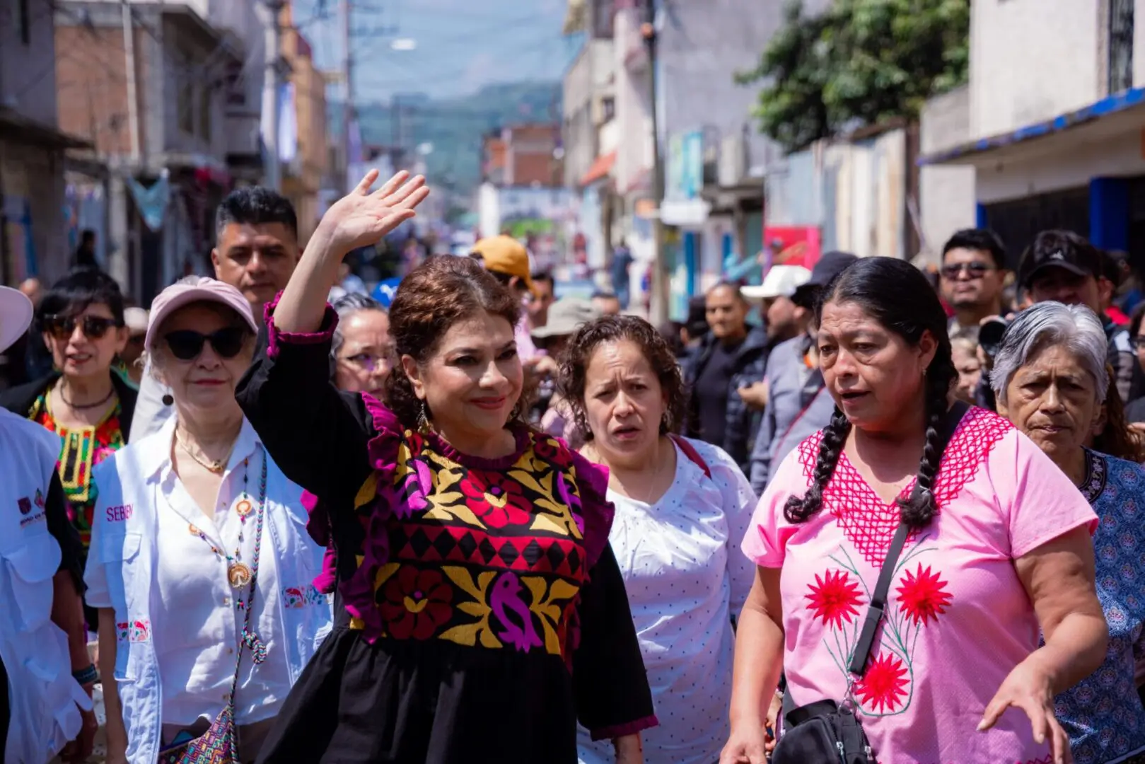 Clara Brugada saluda durante un recorrido comunitario por calles de Xochimilco, acompañada de mujeres con vestimenta tradicional.