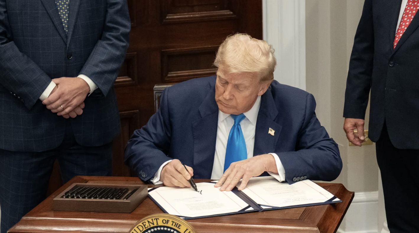 Donald Trump firmando documento en oficina presidencial con bandera de Estados Unidos al fondo