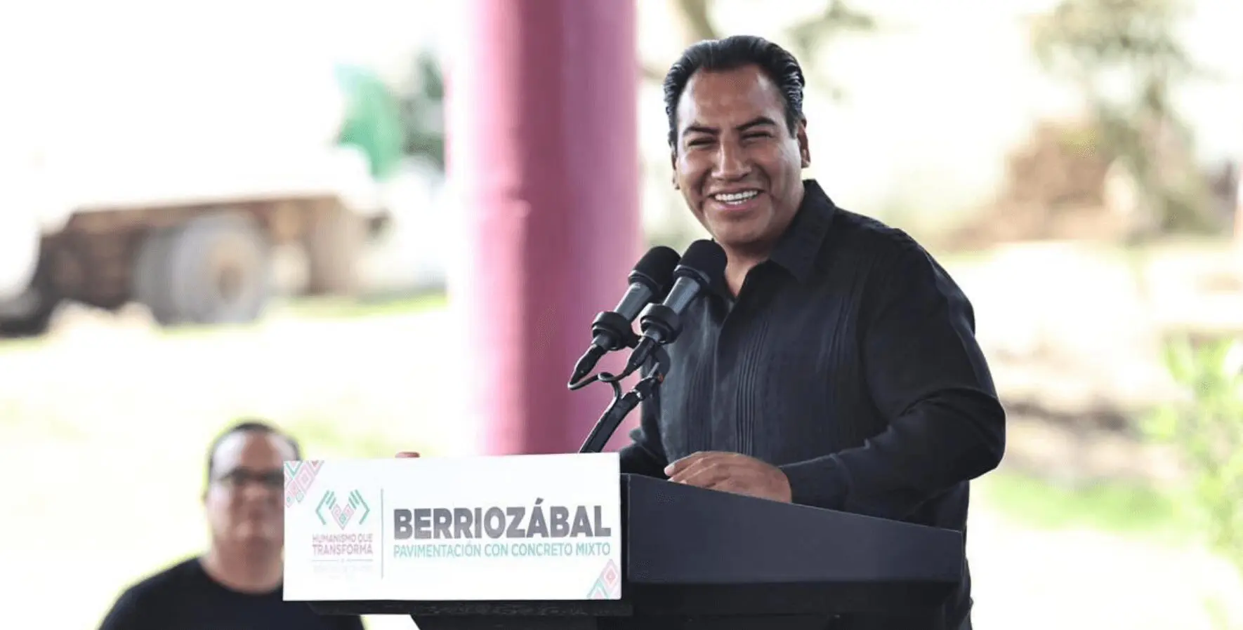 Smiling man speaking at outdoor podium event.