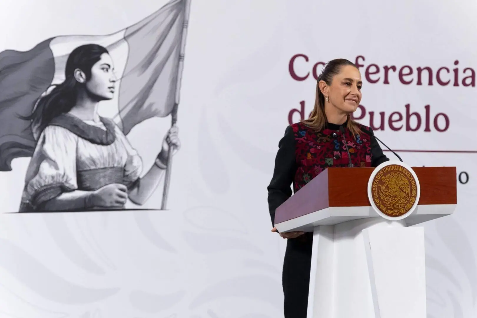 Woman speaking at podium with flag mural