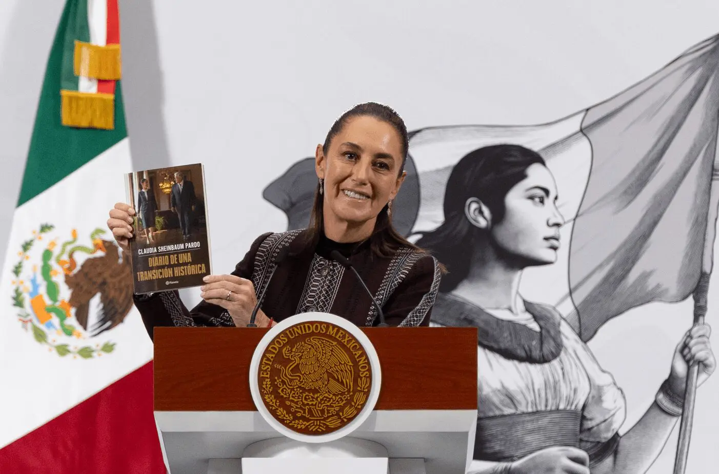 Smiling woman at podium holding book, Mexican flag