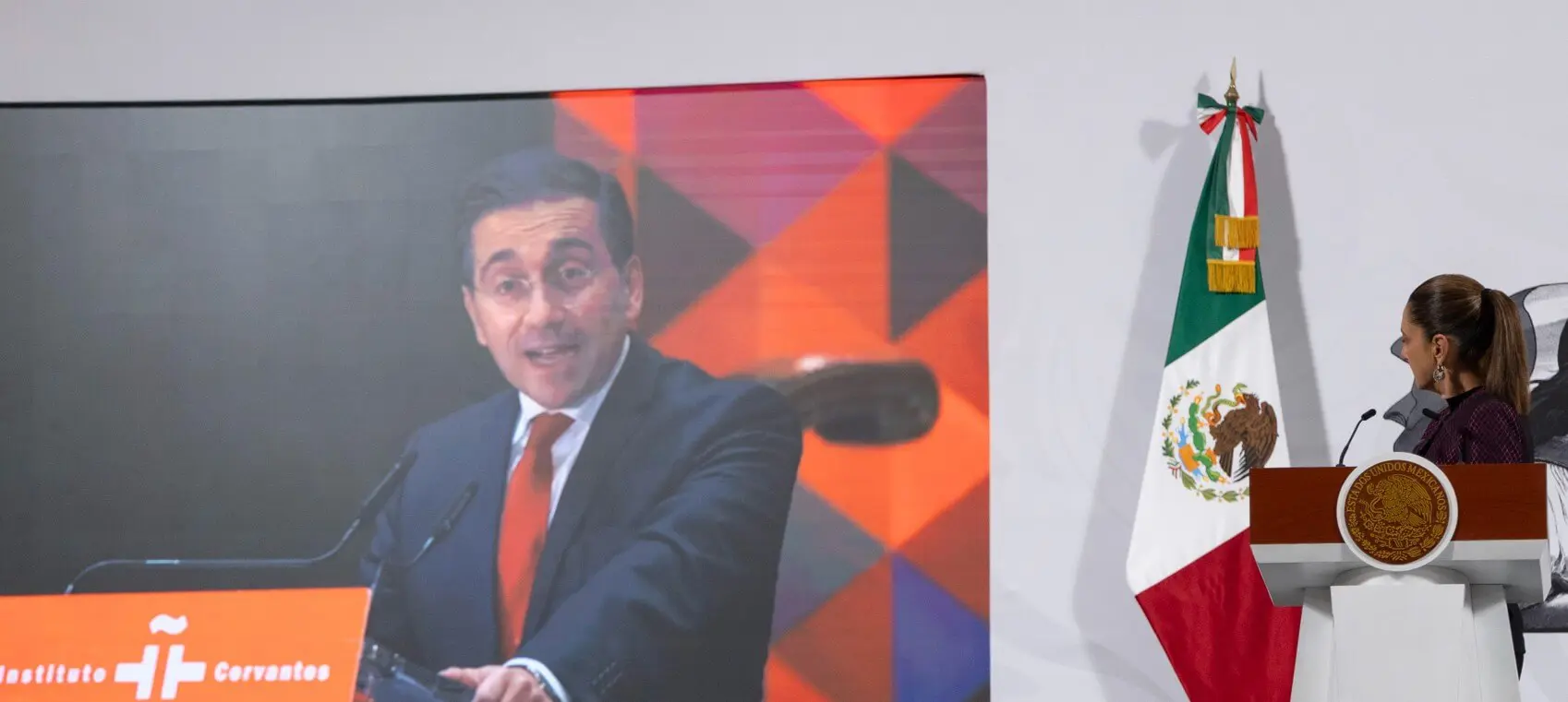 Woman at podium watches screen with Mexican flag