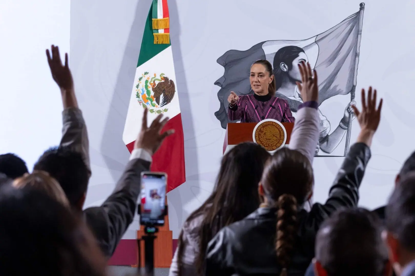 Woman at podium addressing audience with Mexican flag