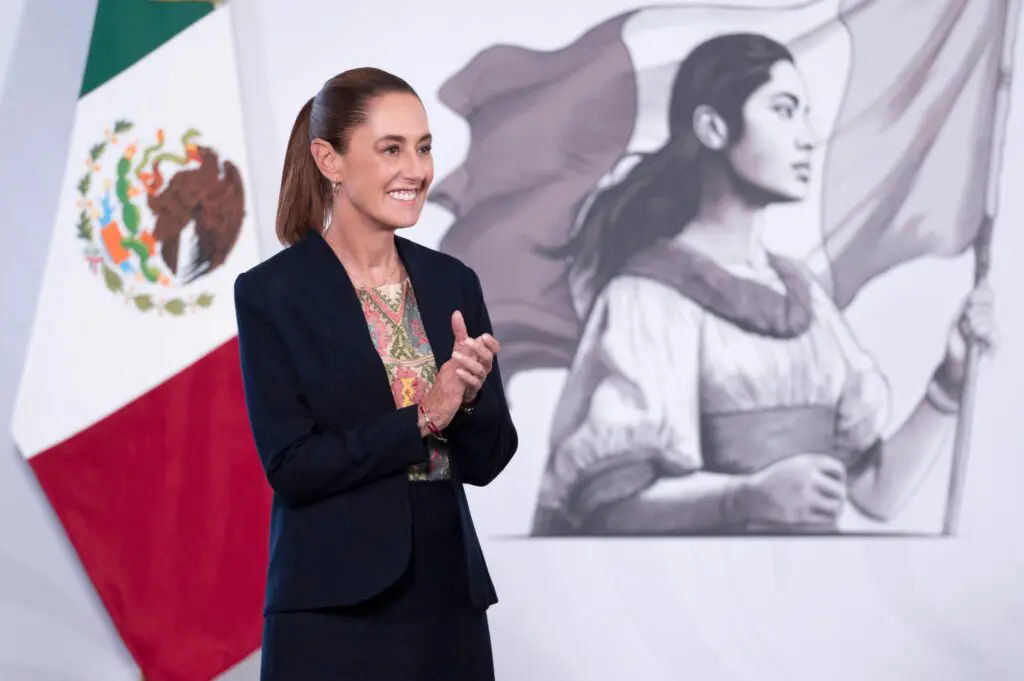 Claudia Sheinbaum durante conferencia matutina en Palacio Nacional, con bandera de México y mural de mujer histórica al fondo.