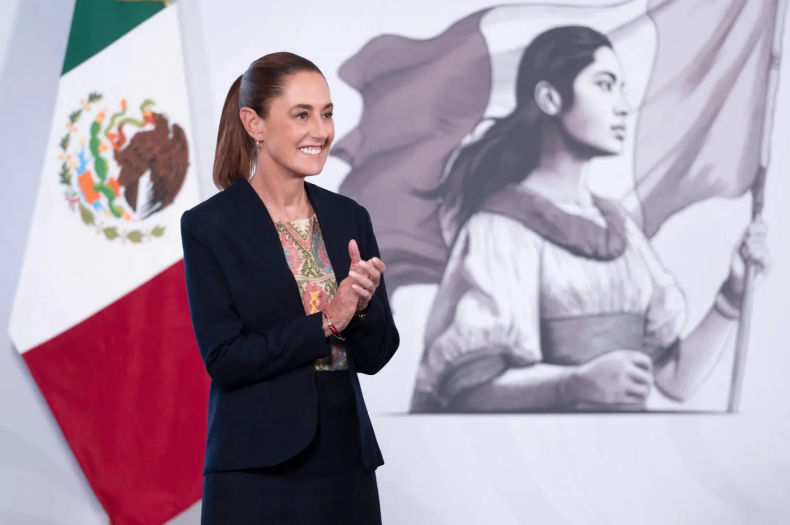 Claudia Sheinbaum durante conferencia matutina en Palacio Nacional, con bandera de México y mural de mujer histórica al fondo.