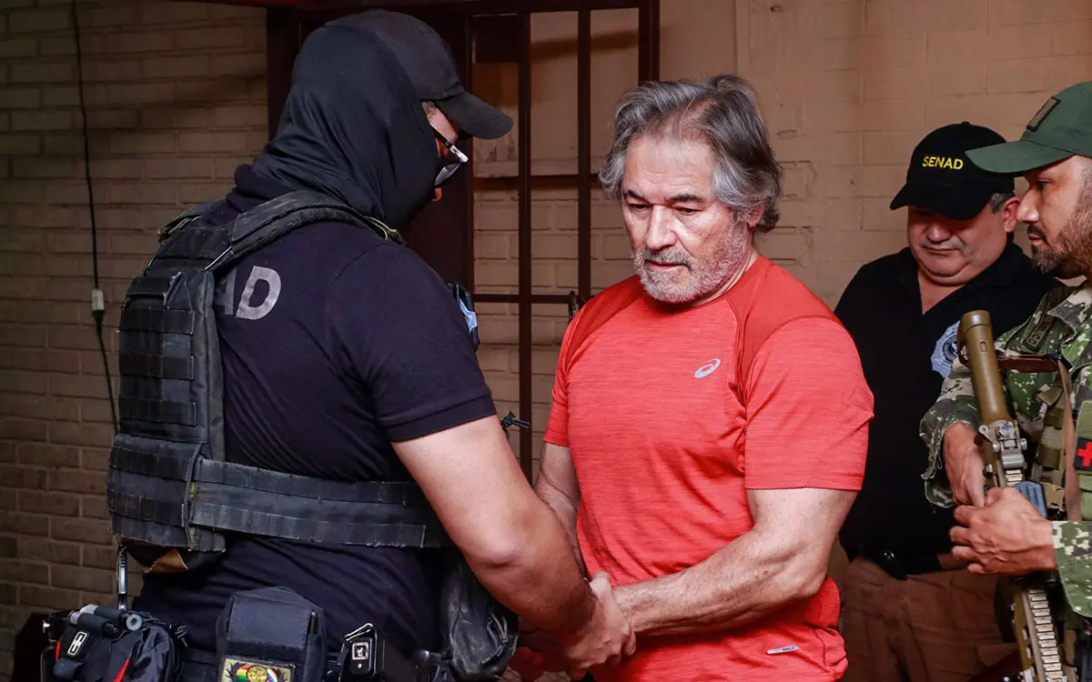 Hernán Bermúdez Requena con uniforme de reo esposado por un elemento de seguridad