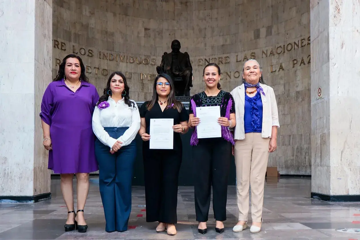 Five women posing indoors, two holding documents