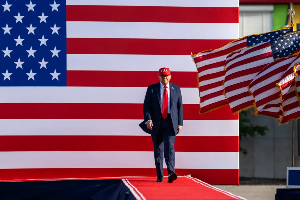 Donald Trump frente a una gran bandera de Estados Unidos con una bandera ondeando en un asta al lado