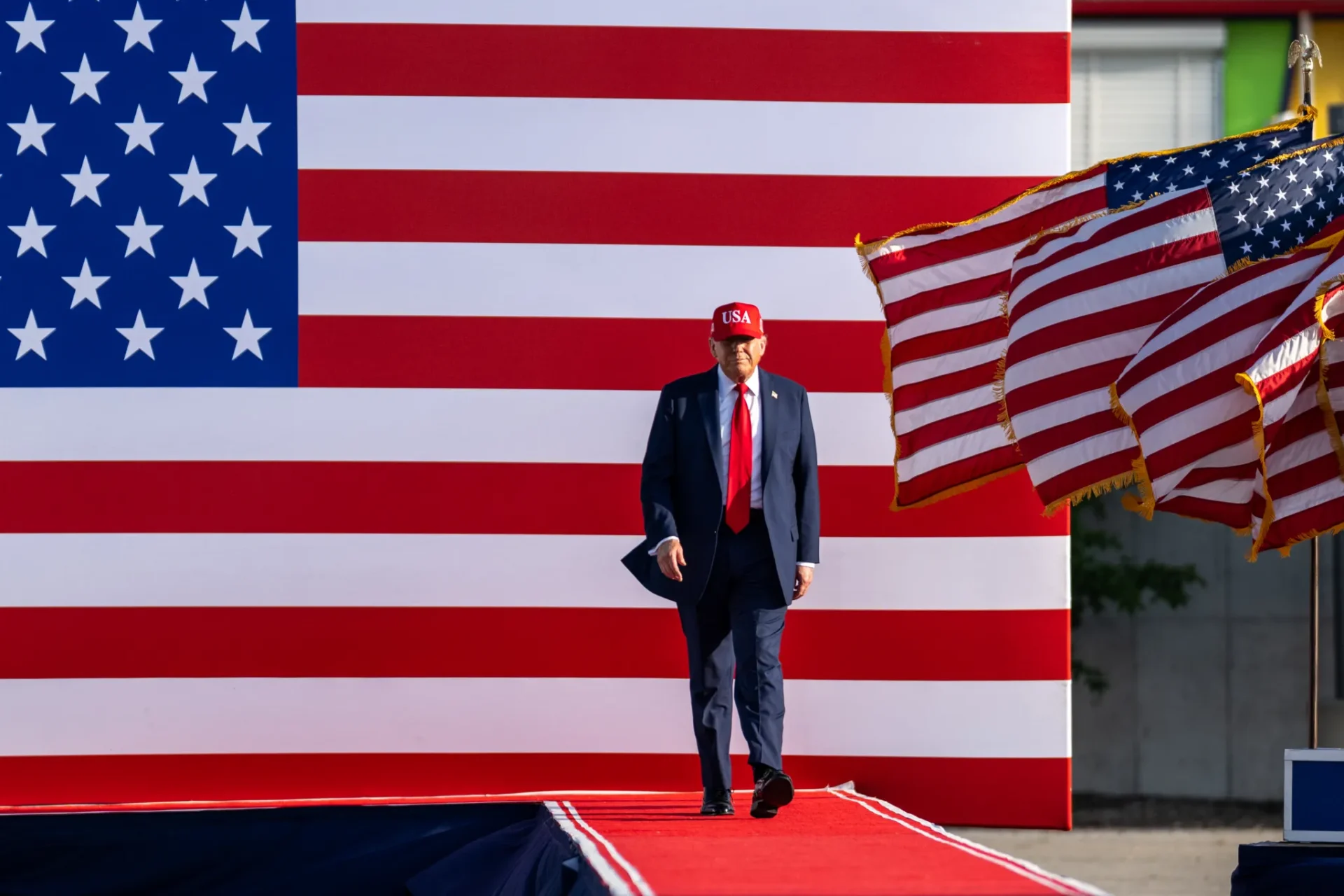 Donald Trump frente a una gran bandera de Estados Unidos con una bandera ondeando en un asta al lado